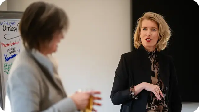 Zwei Frauen unterhalten sich in einer Büroumgebung und veranschaulichen dabei Werte und Haltung im Umgang mit Menschen und in der Kommunikation. Die eine spricht und gestikuliert, während die andere aufmerksam zuhört und ein Glas in der Hand hält. Im Hintergrund ist ein Whiteboard mit bunten Notizen zu sehen.