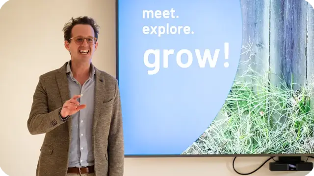 Eine Person in einem Blazer steht lächelnd und gestikulierend vor einer Leinwand, auf der „meet. explore. grow!“ zu lesen ist, vor einem Hintergrund aus Gras und Holzplanken und verkörpert durch ihre einnehmende Ausstrahlung die Werte und Haltung von „mensch & kommunikation“.