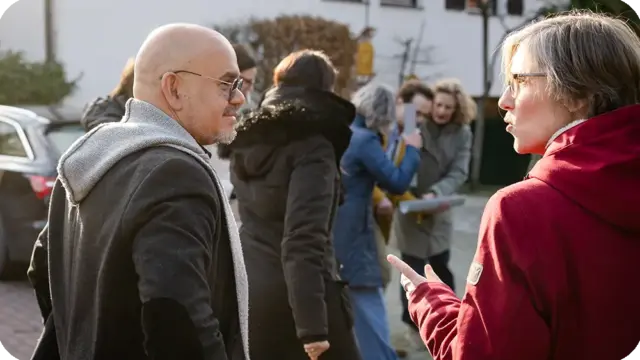 Zwei Personen unterhalten sich im Freien; die eine trägt eine Brille und einen roten Mantel, die andere einen schwarzen Mantel und einen grauen Kapuzenpulli. Ihr Zusammenspiel spiegelt „Werte und Haltung bei Mensch & Kommunikation“ wider, da sie einander aufmerksam zuhören. Im Hintergrund haben sich an diesem sonnigen Tag mehrere weitere Personen versammelt und unterhalten sich.