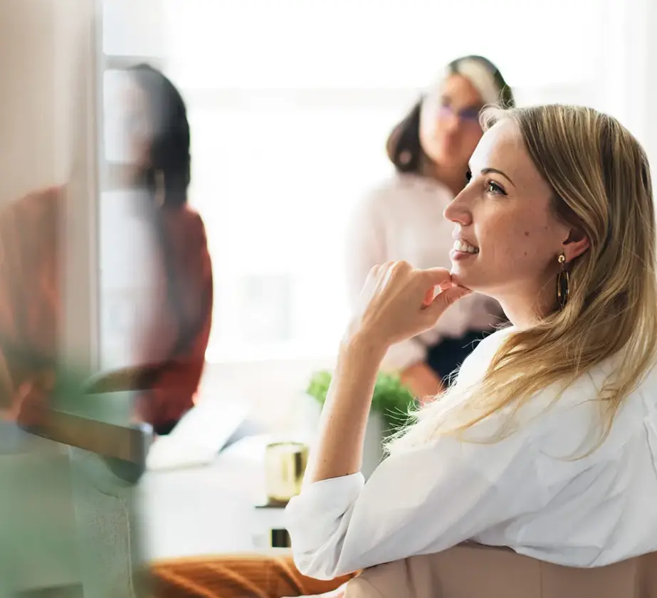 Eine Frau im Vordergrund lächelt und wirkt während einer Besprechung nachdenklich, während im Hintergrund zwei weitere Frauen unscharf zu sehen sind, die in einem hellen Büro sitzen – was verdeutlicht, wie die Verbesserung der Teamarbeit in agilen Strukturen dazu beiträgt, ein positives und produktives Arbeitsumfeld zu schaffen.