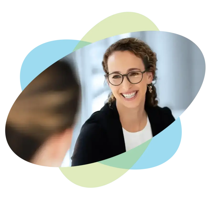 Eine Frau mit lockigem Haar und Brille lächelt, während sie mit einer anderen Person spricht – ein herzlicher Moment im Coaching. Abstrakte blaue und grüne Formen überlagern das Bild, und der Hintergrund ist sanft unscharf.