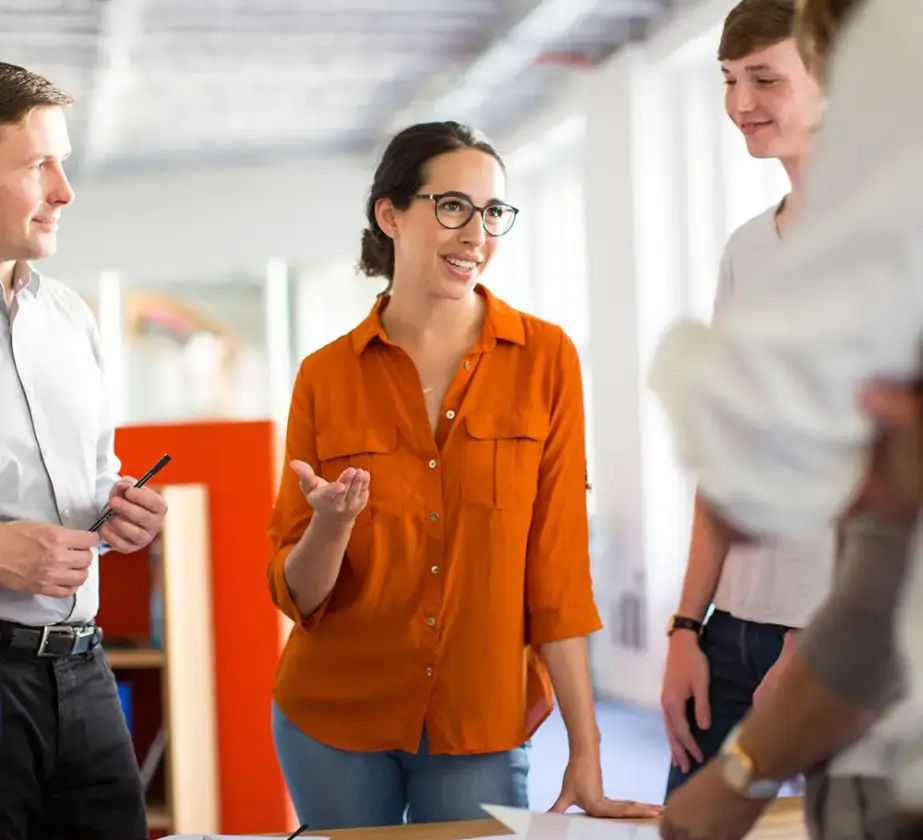 Vier Personen stehen um einen Tisch mit Unterlagen herum und führen ein freundliches Gespräch. Eine Frau in einem orangefarbenen Shirt spricht, während die anderen zuhören und lächeln. Die Szene scheint in einem modernen Büro zu spielen, wo sie sich damit beschäftigen, wie man die Teamarbeit in agilen Strukturen verbessern kann.