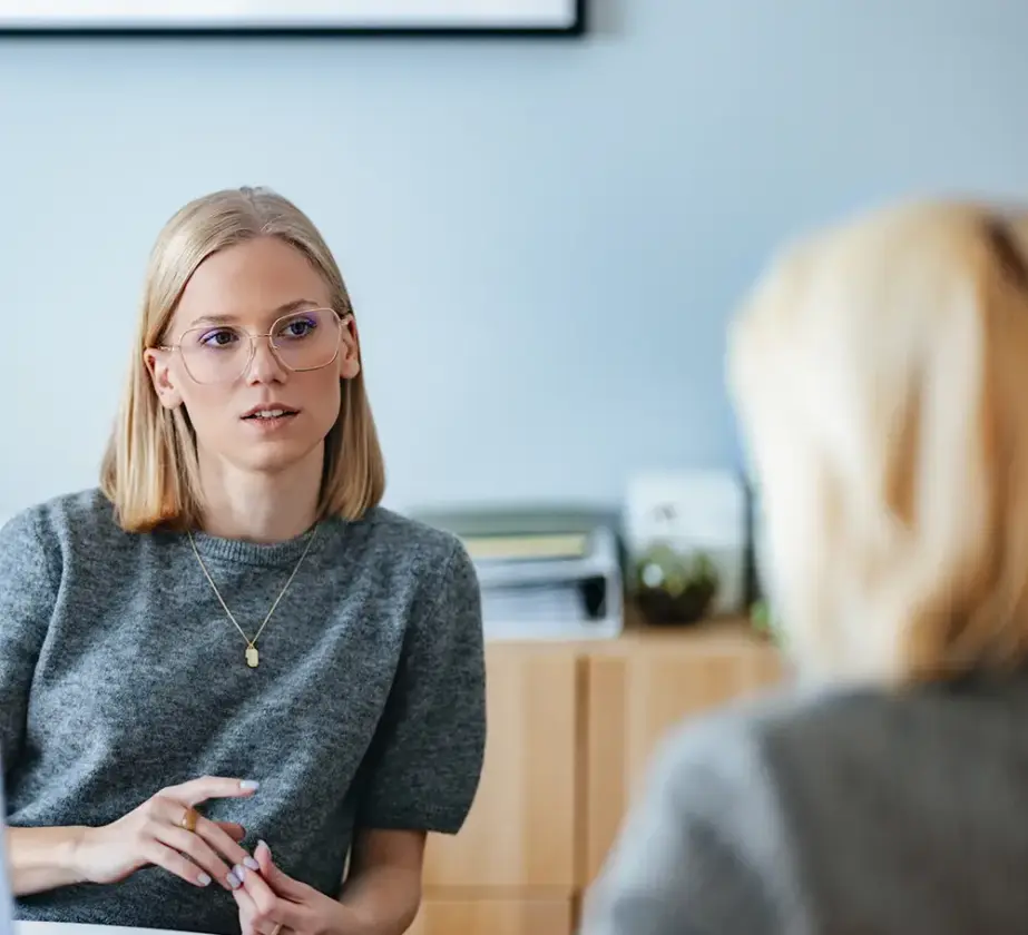 Eine Frau mit blonden Haaren und Brille sitzt an einem Tisch und unterhält sich mit einer anderen Person, die der Kamera den Rücken zuwendet. Der Schauplatz scheint ein Büro zu sein, im Hintergrund sind Regale und Unterlagen zu sehen – eine ideale Umgebung für Gespräche darüber, wie sich die Teamarbeit in agilen Strukturen verbessern lässt.
