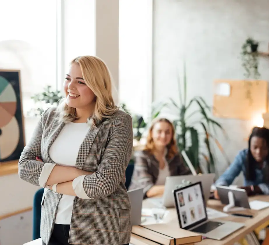 Eine lächelnde Frau in einem Blazer steht mit verschränkten Armen in einem modernen Büro und verkörpert den Geist der „Teamarbeit in agilen Strukturen verbessern“. Zwei Kollegen sitzen an einem Schreibtisch mit Laptops und Unterlagen und arbeiten zusammen, während Pflanzen den hellen Arbeitsbereich schmücken.
