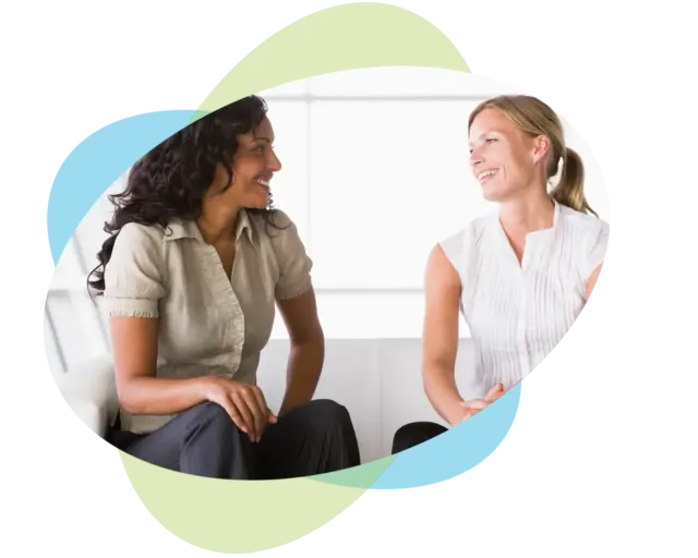 Zwei Frauen sitzen auf einem Sofa, lächeln und unterhalten sich in einem hellen Raum, während abstrakte grüne und blaue Formen das Bild einrahmen – ein Bild, das den Geist von „Weiterbildung für Führung und Kommunikation“ perfekt einfängt.