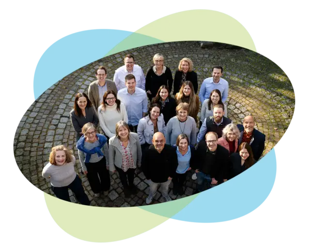 Eine Gruppe von etwa 24 Erwachsenen steht gemeinsam im Freien auf einem kreisförmigen Steinweg, blickt nach oben und lächelt in die Kamera – vielleicht haben sie sich zu einem Führungstraining für Führungskräfte versammelt. Im Hintergrund sind sich überlappende blaue und grüne ovale Formen zu sehen.