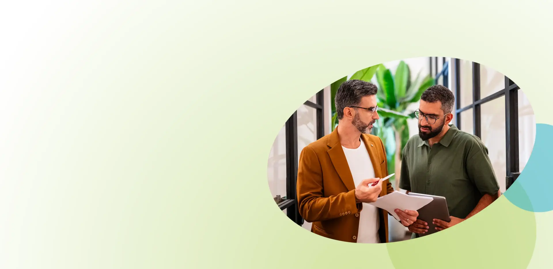 Zwei Männer stehen zusammen in einem Raum und besprechen während eines Konfliktmanagement-Seminars für Unternehmen einige Unterlagen. Der eine trägt einen braunen Blazer und eine Brille und hält einen Stift in der Hand; der andere trägt ein grünes Hemd und hält Unterlagen in der Hand. Im Hintergrund sind Grünpflanzen zu sehen.