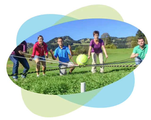 Fünf Personen arbeiten im Freien gemeinsam daran, mithilfe von Seilen und Stangen das Gleichgewicht zu halten und einen großen Tennisball über eine Wiese zu bewegen, während im Hintergrund Hügel und Bäume vor einem blauen Himmel zu sehen sind – eine spannende Übung, die häufig im Rhetoriktraining für Fach- und Führungskräfte eingesetzt wird, um die Kommunikations- und Teamfähigkeiten zu verbessern.