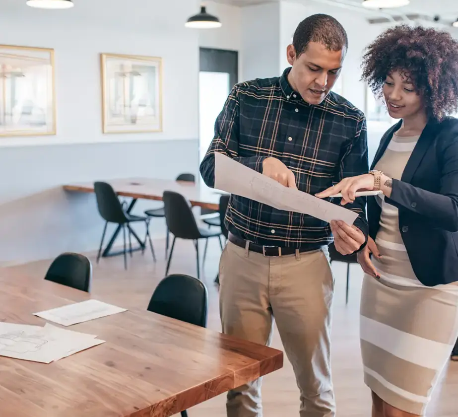 Zwei Personen stehen in einem modernen Büro mit Holztischen und -stühlen und besprechen ein Dokument; sie zeigen auf das Papier und unterhalten sich darüber, wie sie die Teamarbeit in agilen Strukturen verbessern können.