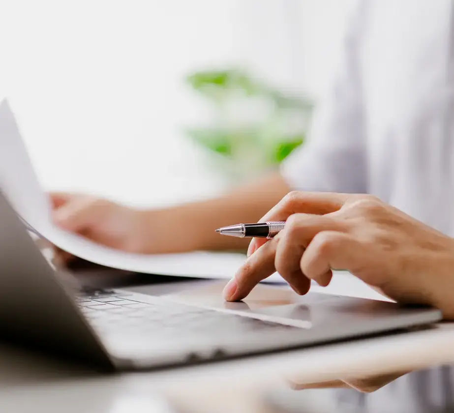 Eine Person, die einen Stift in der Hand hält, arbeitet am Laptop und sieht sich dabei ein Dokument an, im Hintergrund ist eine verschwommene grüne Pflanze zu sehen – eine ideale Kulisse, um die Teamarbeit in agilen Strukturen zu verbessern.