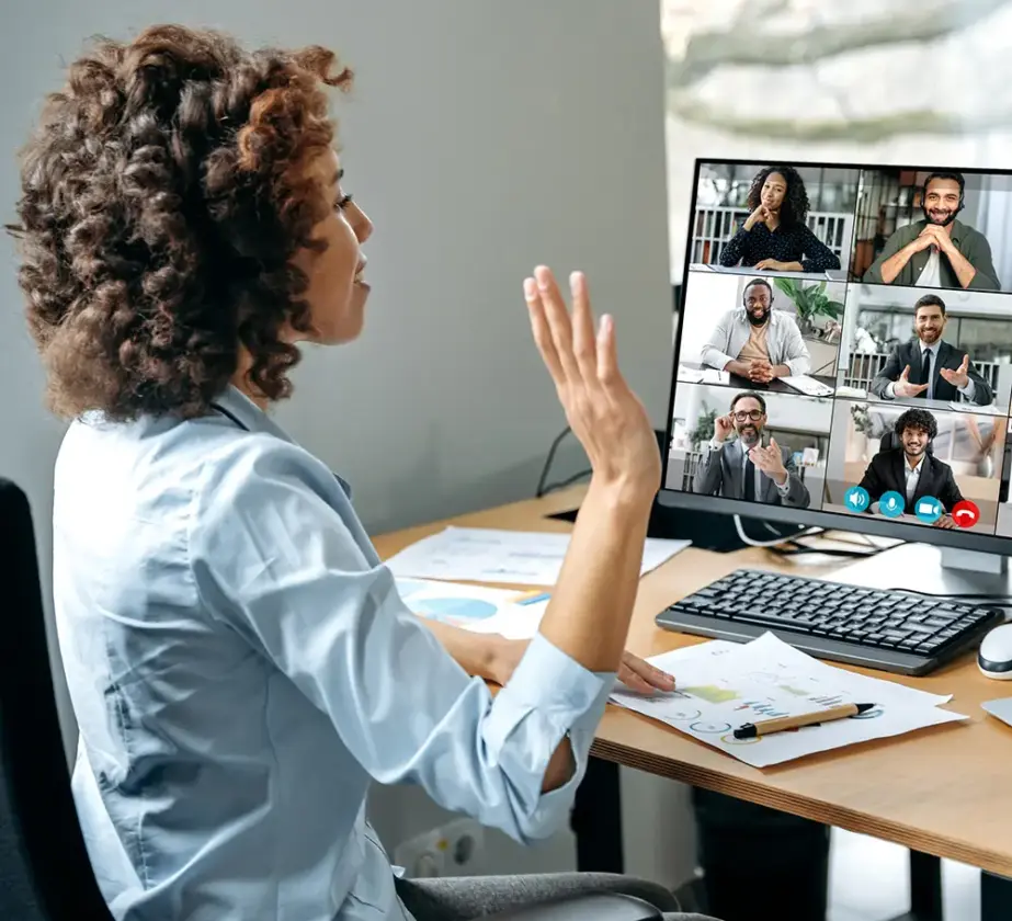 Eine Frau sitzt an einem Schreibtisch und hebt die Hand, während sie an einer Videokonferenz mit sechs Personen teilnimmt, die auf ihrem Computerbildschirm zu sehen sind. Auf dem Schreibtisch liegen Unterlagen und eine Tastatur, während sie sich aktiv an der Diskussion darüber beteiligt, wie man die Teamarbeit in agilen Strukturen verbessern kann.