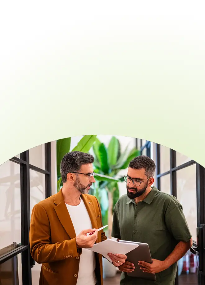 Zwei Männer in einem modernen Büro besprechen Dokumente für ein Konfliktmanagement-Seminar für Unternehmen, während sie ein Tablet in der Hand halten.