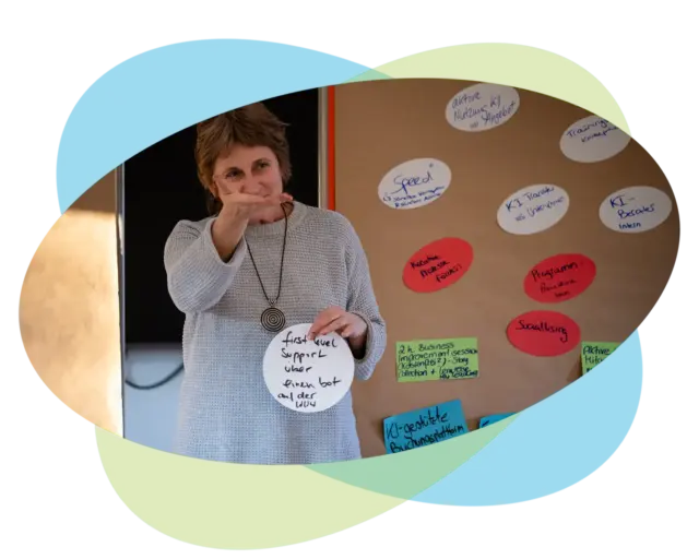 Eine Frau hält ein rundes Schild vor einer Pinnwand, die mit Haftnotizen bedeckt ist, auf denen deutsche und englische Projektbegriffe für Trainingslösungen mit Wirkung stehen.
